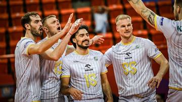 Grupa siatkarzy w biało-ż&oacute;łtych strojach świętuje zwycięstwo, jeden z zawodnik&oacute;w unosi rękę w geście triumfu.