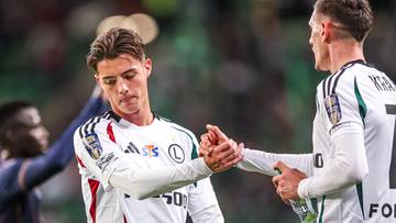 Dw&oacute;ch piłkarzy Legii Warszawa w białych strojach sportowych ściska sobie dłonie na boisku.