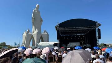Zgromadzenie ludzi pod parasolami przed dużą figurą religijną i sceną polową w słoneczny dzień.