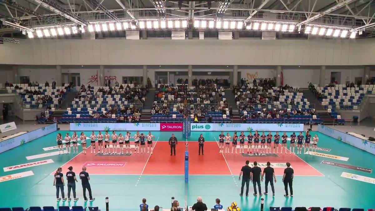 Dwie drużyny siatkarek stoją po obu stronach siatki na boisku w hali sportowej.