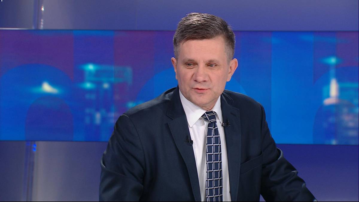 Mężczyzna w granatowym garniturze i krawacie siedzi przy stole przed niebieskim tłem.