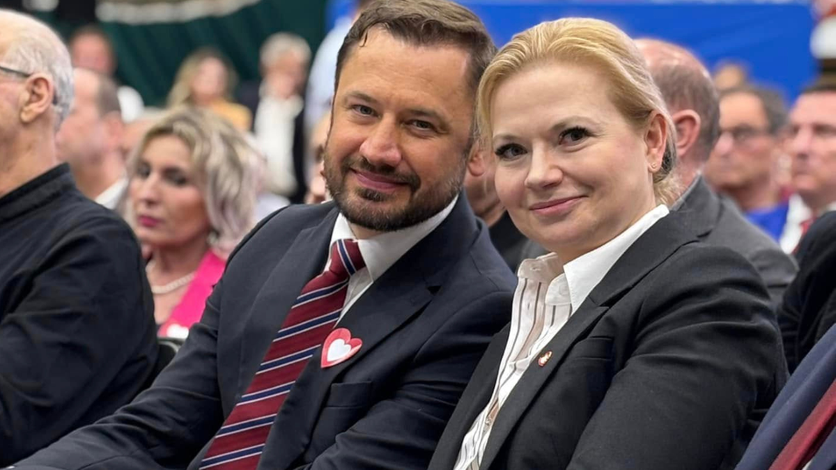 Uśmiechnięta para polityk&oacute;w siedzi w pierwszym rzędzie na wydarzeniu.