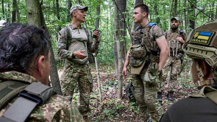 Grupa żołnierzy w mundurach polowych w lesie. Dw&oacute;ch żołnierzy rozmawia, jeden trzyma w ręku kr&oacute;tkofal&oacute;wkę. Na rękawie jednego z nich widoczna naszywka z flagą Ukrainy.