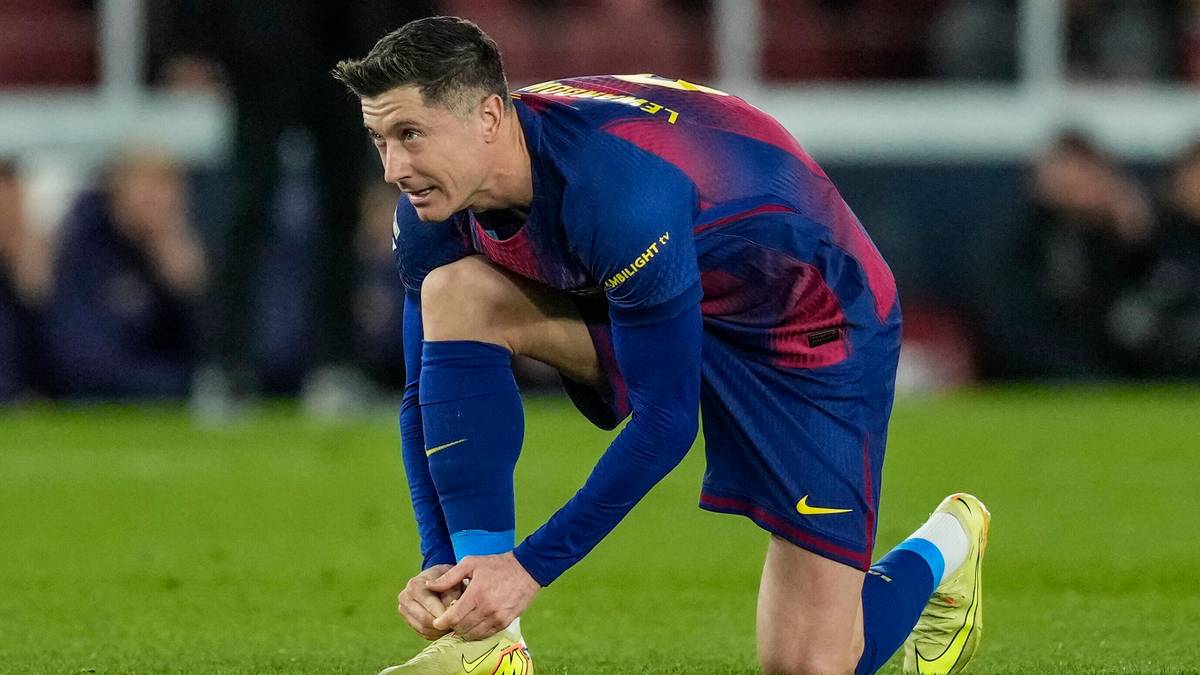 Robert Lewandowski w stroju FC Barcelony klęczy na boisku i zawiązuje sznurówki butów.