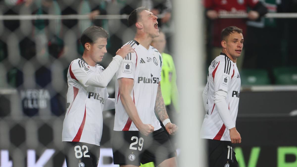 Trzech piłkarzy Legii Warszawa w białych strojach z czarnymi detalami, jeden z nich świętuje strzelenie bramki z uniesionymi rękami i otwartymi ustami.