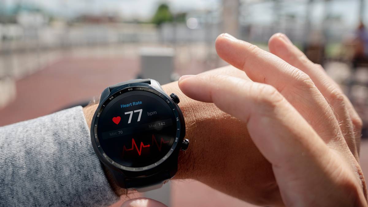 Zbliżenie na nadgarstek osoby noszącej smartwatch. Na ekranie zegarka widoczny jest symbol serca i aktualny puls wynoszący 77 uderzeń na minutę, wraz z wykresem EKG.