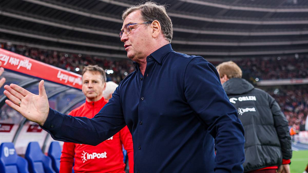 Trener piłkarski w okularach i granatowej koszuli, gestykuluje prawą ręką na tle stadionu.