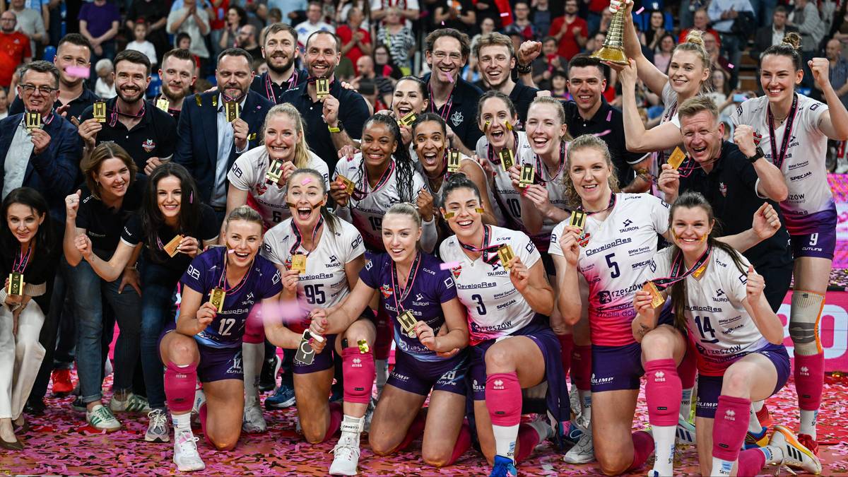 Drużyna siatkarek w strojach sportowych pozuje do zdjęcia z trofeum i medalami, ciesząc się ze zwycięstwa. W tle widownia.