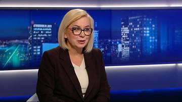 Kobieta w okularach i ciemnym żakecie wypowiada się w studio telewizyjnym na tle nocnej panoramy miasta.