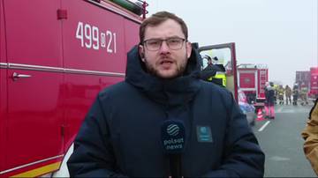 Mężczyzna w okularach i ciemnej kurtce stoi przed czerwonym wozem strażackim, trzymając mikrofon z logo Polsat News. Za nim widać innych strażaków i pojazdy.