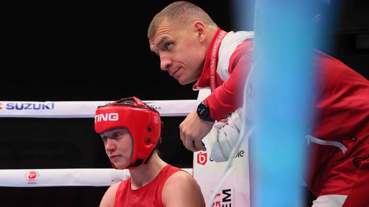 Trener spogląda na młodą pięściarkę w czerwonym kasku ochronnym, siedzącą przy ringu bokserskim.