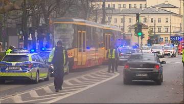 Widok na miejsce wypadku drogowego w Warszawie. Widać uszkodzony tramwaj, autobus, radiowozy policyjne z włączonymi niebieskimi sygnałami świetlnymi oraz inne pojazdy. Na ulicy obecni są ludzie w kamizelkach odblaskowych.
