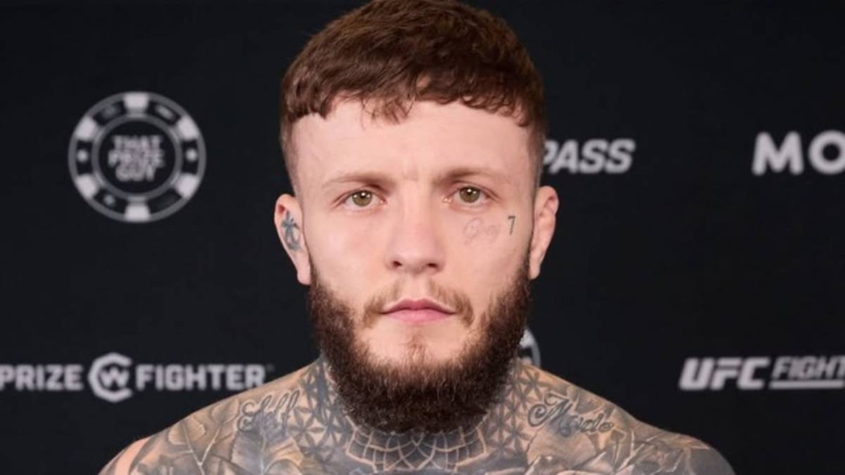 Mężczyzna z tatuażami na twarzy i ciele, krótkimi włosami i brodą, patrzący na wprost.