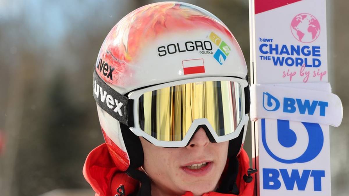 Skoczek narciarski w białym kasku z kolorowymi akcentami i złotymi goglami.