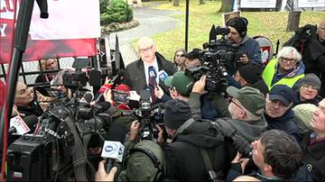 Bogdan Święczkowski otoczony przez dziennikarzy z mikrofonami i kamerami podczas protestu.