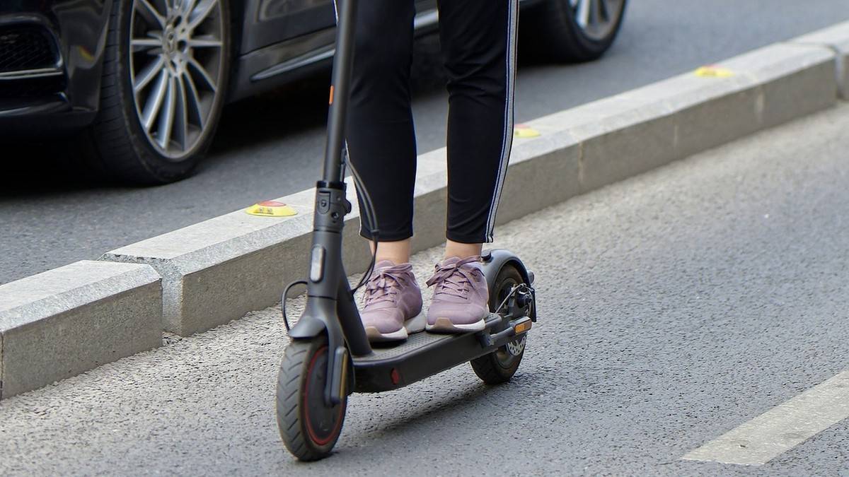 Osoba jadąca na hulajnogie elektrycznej przy krawężniku drogi.