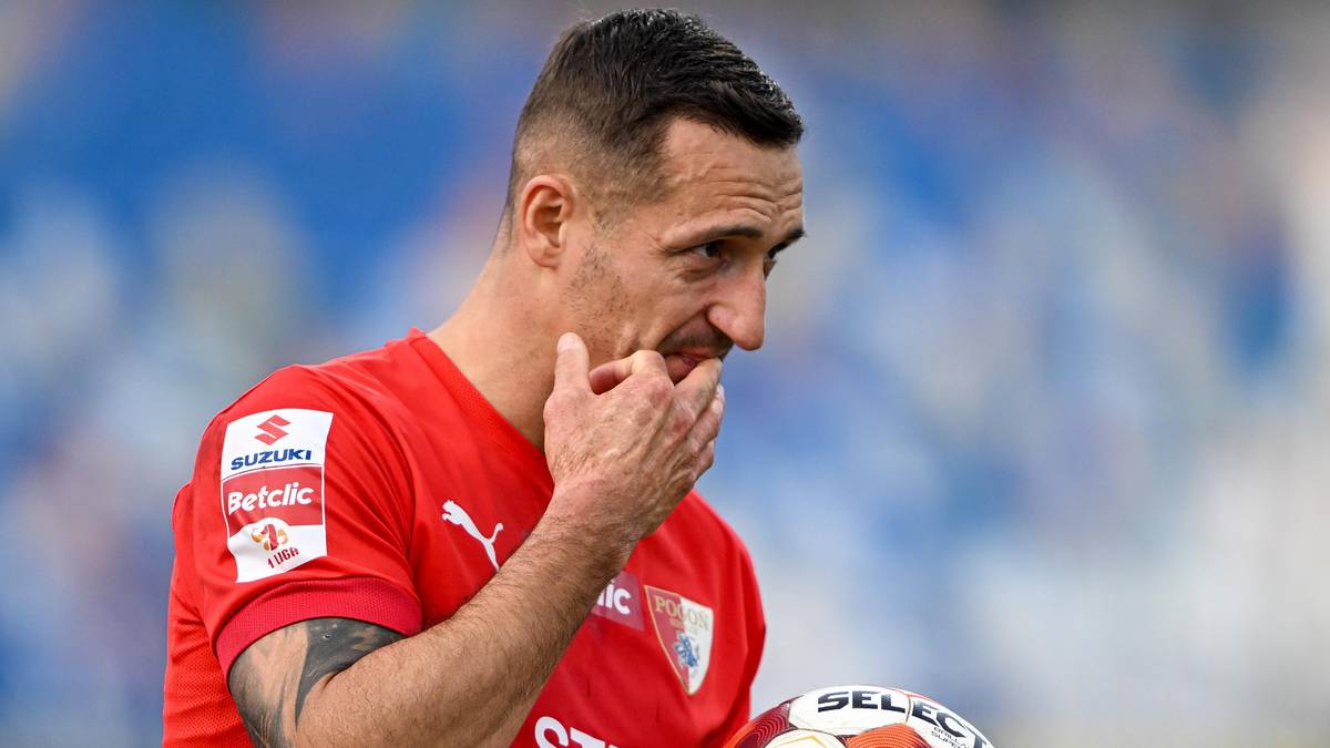 Piłkarz w czerwonej koszulce trzyma piłkę i zastanawia się.