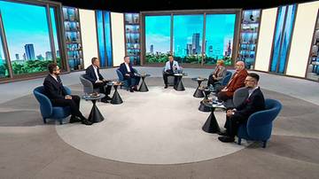 Siedem mężczyzn w garniturach siedzi w okręgu na fotelach w studiu telewizyjnym, uczestnicząc w dyskusji.