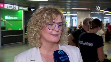 Otylia Jędrzejczak z kręconymi blond włosami i okularami w niebieskich oprawkach, ubrana w białą marynarkę, trzyma mikrofon z logo Polsat Sport. W tle widać ludzi i elementy hali lotniskowej z zielonymi neonami.