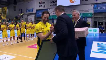 Grzegorz Łomacz, siatkarz w ż&oacute;łtej koszulce, odbiera ramkę z dyplomem od mężczyzny w garniturze. Obok stoi drugi mężczyzna z pudełkiem prezentowym. W tle widać innych sportowc&oacute;w i reklamy.