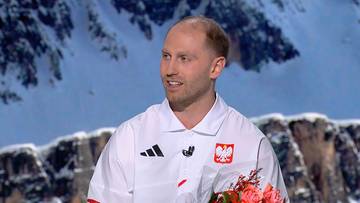 Damian Żurek, ubrany w białą koszulkę sportową z godłem Polski, pozuje z bukietem kwiatów na tle zaśnieżonych gór.