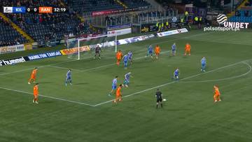 Mecz piłki nożnej pomiędzy drużynami Kilmarnock FC i Rangers FC. Zawodnicy w pomarańczowych i niebieskich strojach grają na zielonej murawie stadionu. Wynik meczu na ekranie: KIL 0 - 0 RAN, czas 32:19.