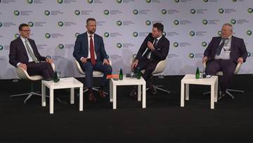 Czterech mężczyzn siedzi na krzesłach za białymi stolikami przed ścianą z logo.