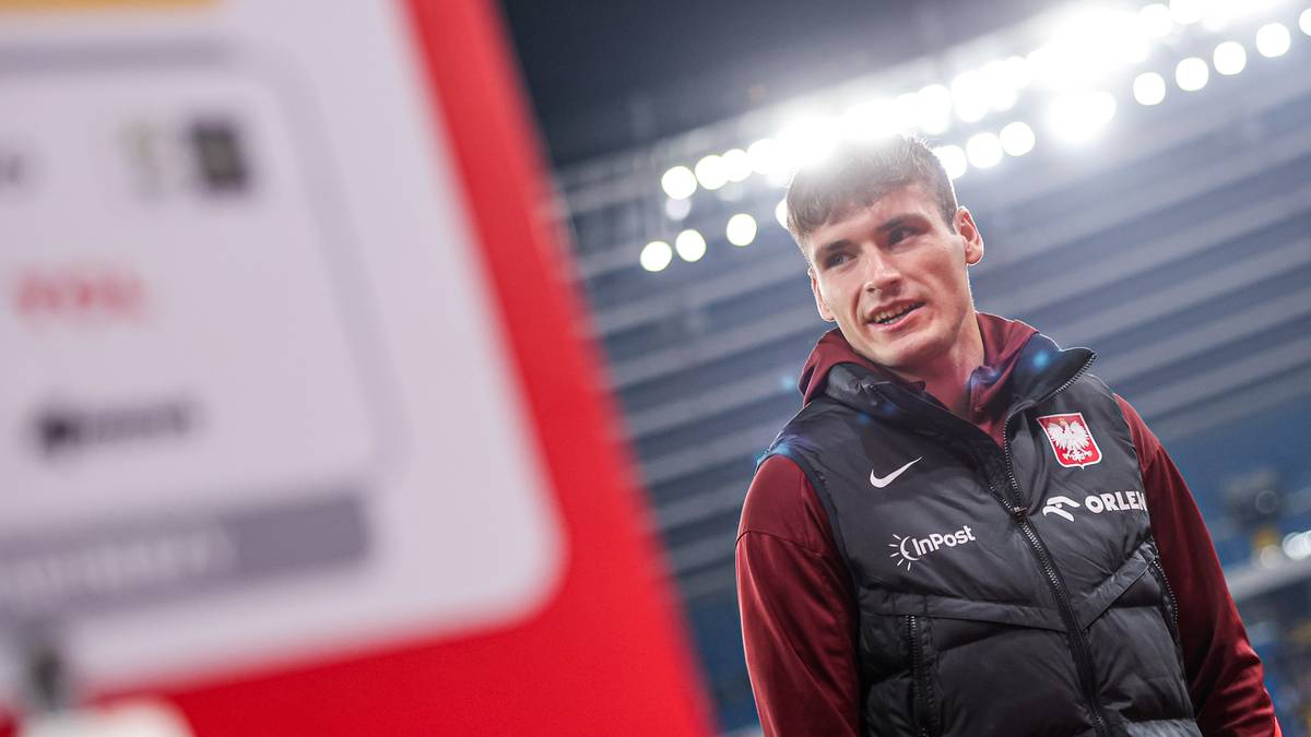 Młody mężczyzna w stroju sportowym, prawdopodobnie piłkarz, w ciemnej kamizelce z herbem Polski na piersi, patrzy w bok z uśmiechem. W tle widać stadion i światła.