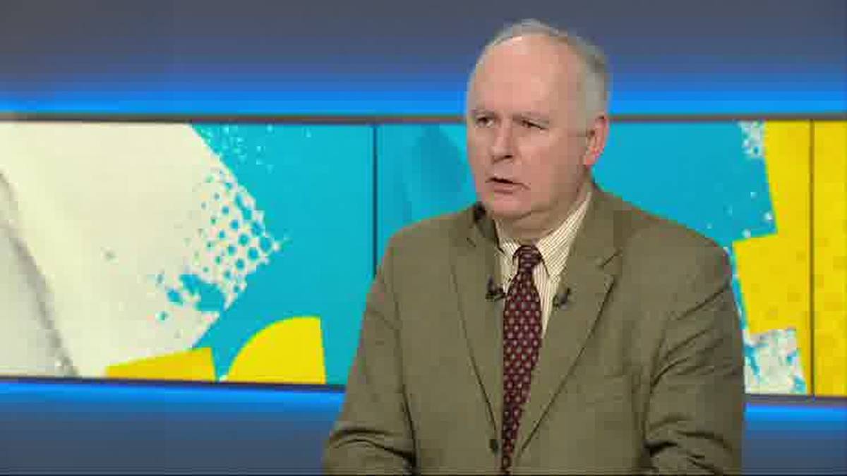 Mężczyzna w jasnobrązowym garniturze i krawacie z wzorem, siedzący na tle niebiesko-ż&oacute;łtego, abstrakcyjnego wzoru na ekranie.