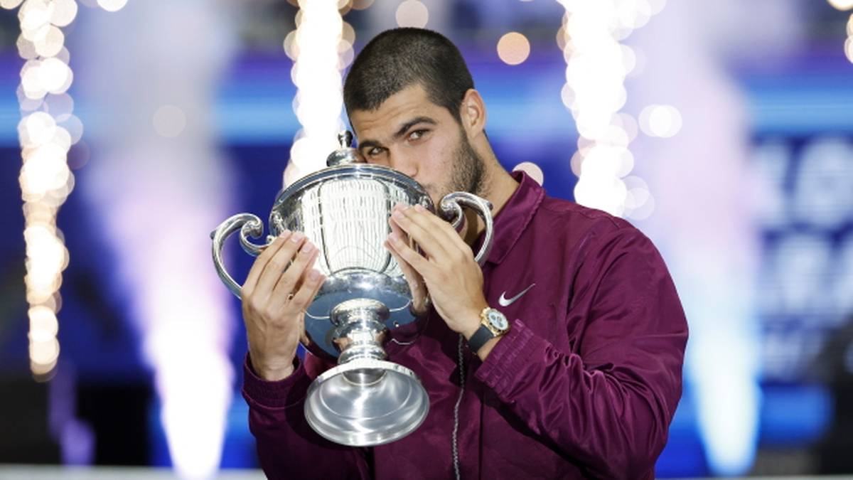 Carlos Alcaraz całuje puchar tenisowy na podium po wygranej. Jest ubrany w bordową kurtkę sportową z zegarkiem na ręce.