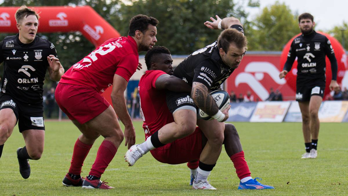 Dwóch graczy w czerwonych strojach próbuje powstrzymać gracza w czarnym stroju, który próbuje zdobyć przyłożenie podczas meczu rugby.