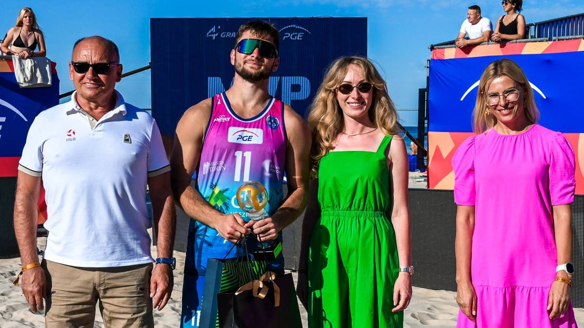 Cztery osoby pozujące do zdjęcia na plaży, dwie pośrodku w strojach sportowych i sukience, dwie po bokach w strojach casualowych.