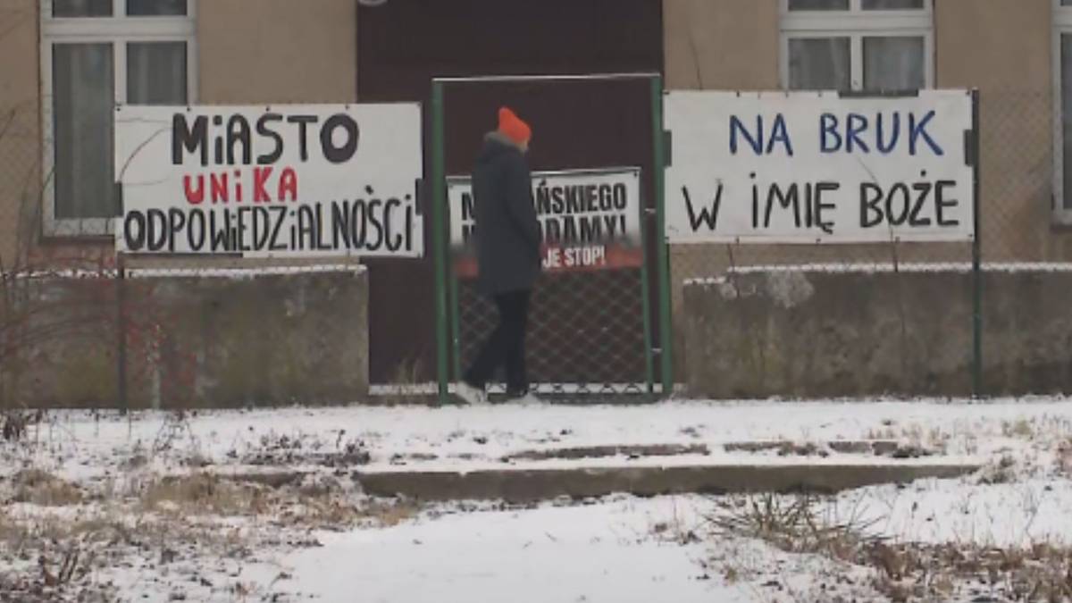 Trzy tablice z napisami "MIASTO UNIKA ODPOWIEDZIALNOŚCI", "MIEJSKIEGO DAMY JE ST" oraz "NA BRUK W IMIĘ BOŻE" przed budynkiem, na tle zaśnieżonego otoczenia.