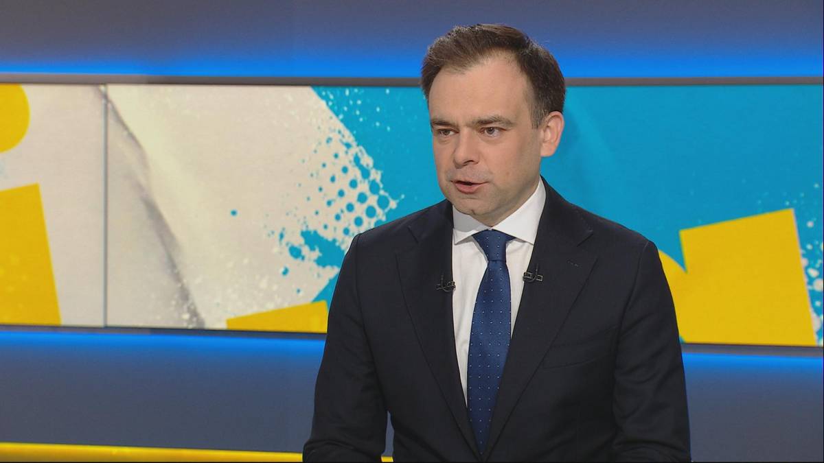 Mężczyzna w garniturze i krawacie przemawia przed kolorowym tłem.