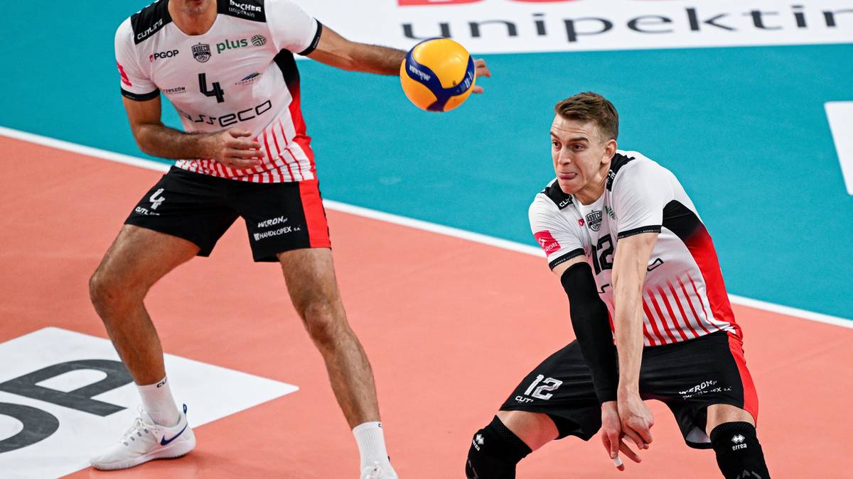 Dwóch siatkarzy w białych koszulkach z czerwonymi elementami i czarnych spodenkach na boisku siatkarskim.