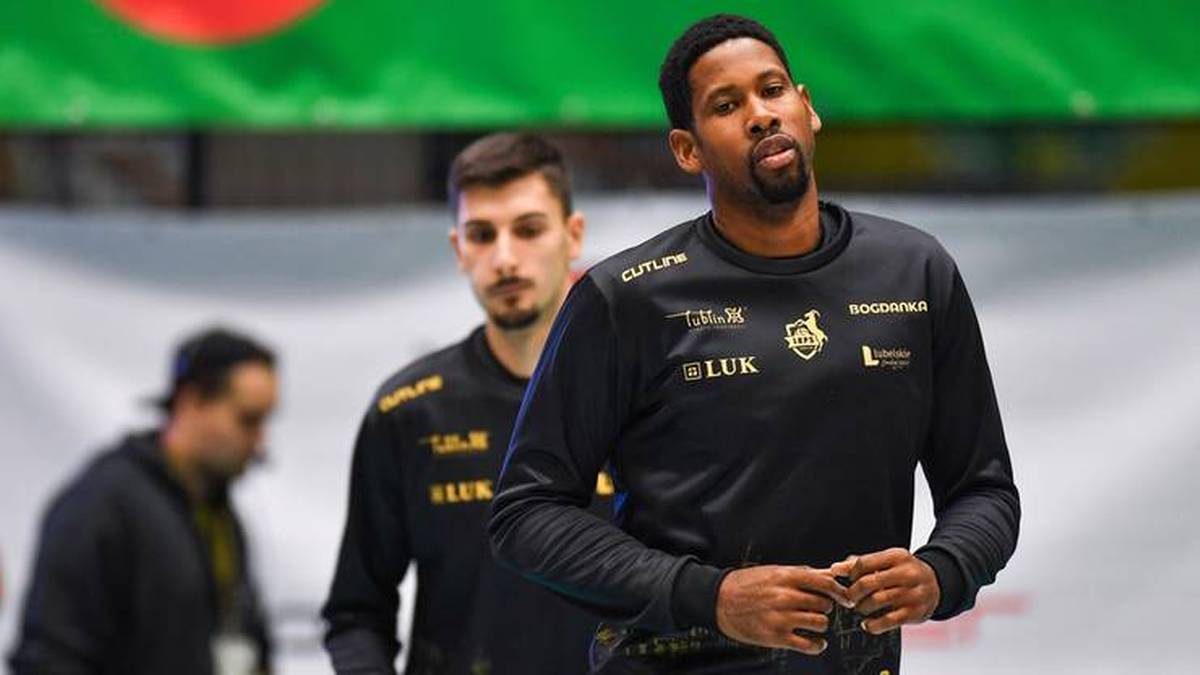 Trzech mężczyzn w strojach sportowych, na pierwszym planie czarnoskóry mężczyzna w czarnej bluzie z napisami, z tyłu dwaj biali mężczyźni w podobnych bluzach.