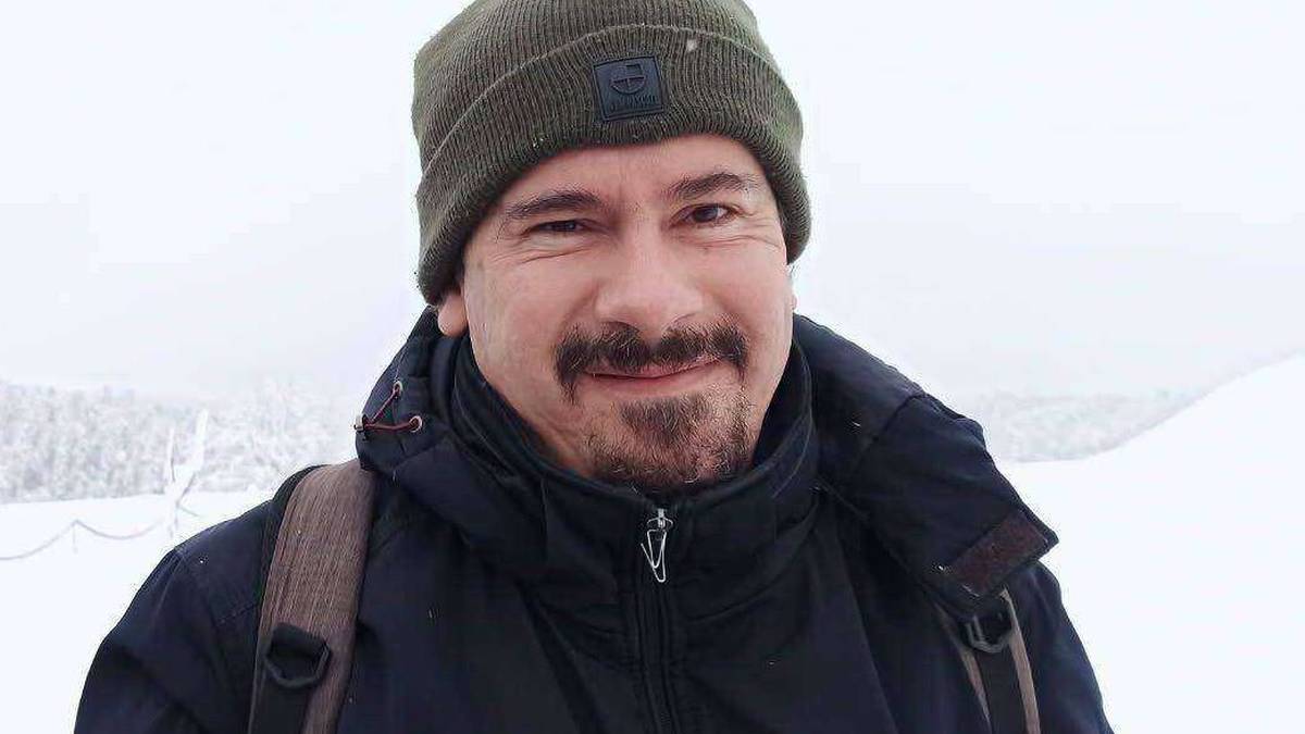 Mężczyzna w zimowej kurtce i czapce z plecakiem na ramionach, uśmiecha się na tle zimowego krajobrazu.