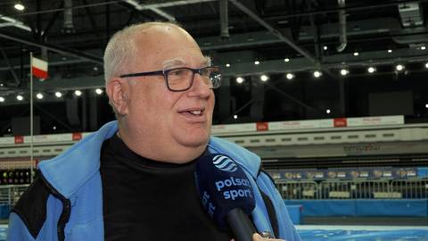 Mężczyzna w okularach trzyma mikrofon, na tle hali sportowej.