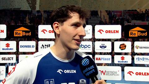 Młody mężczyzna w stroju sportowym, uśmiechnięty, trzymający mikrofon, na tle tablic z logotypami sponsorów.