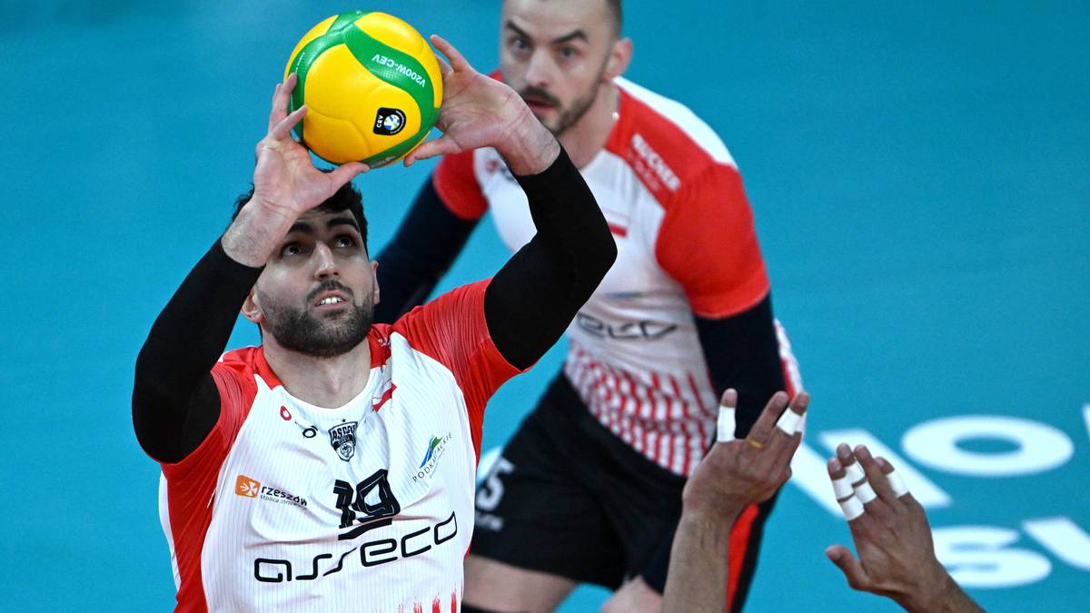 Siatkarz w białej koszulce z czerwonymi pasami i logo Asseco Resovii, przygotowuje się do zagrywki, trzymając piłkę siatkową.