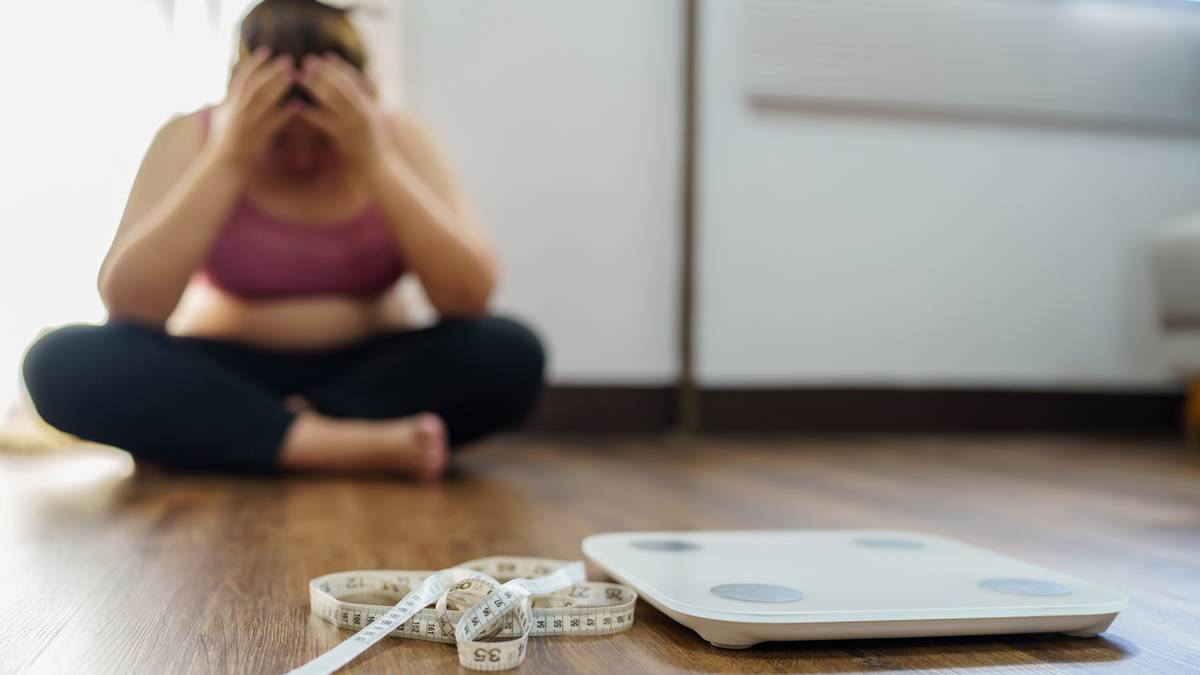 Kobieta siedzi na podłodze, zakrywając twarz dłońmi, obok niej leży waga łazienkowa i taśma miernicza.