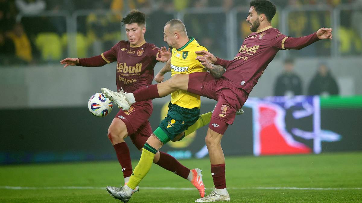 Trzech piłkarzy walczy o piłkę na boisku, dw&oacute;ch w bordowych strojach i jeden w ż&oacute;łtym.