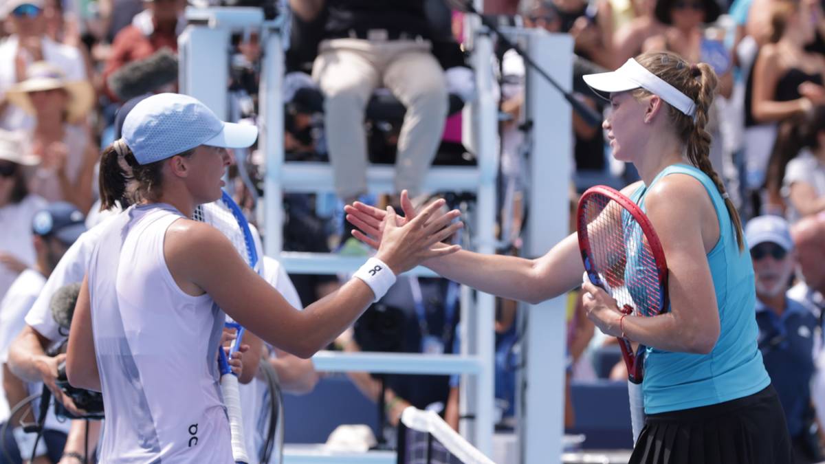 Dwie tenisistki po meczu na korcie, podają sobie dłonie w geście gratulacji. Jedna z nich w białej czapeczce, druga w niebieskiej koszulce i białej czapce z daszkiem.