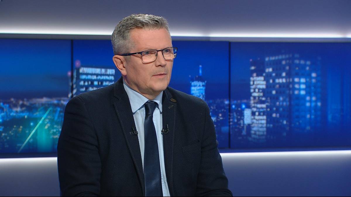 Mężczyzna w okularach i garniturze siedzi przed ekranem z widokiem nocnego miasta, wypowiadając się.