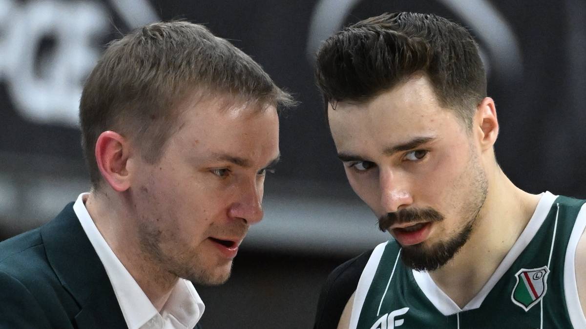 Dwóch mężczyzn w strojach sportowych rozmawia, jeden trener w garniturze i drugi zawodnik w koszulce sportowej.