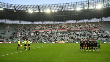Boisko piłkarskie ze stadionem i widzami na trybunach, z sędziami po jednej stronie i drużyną piłkarzy po drugiej.