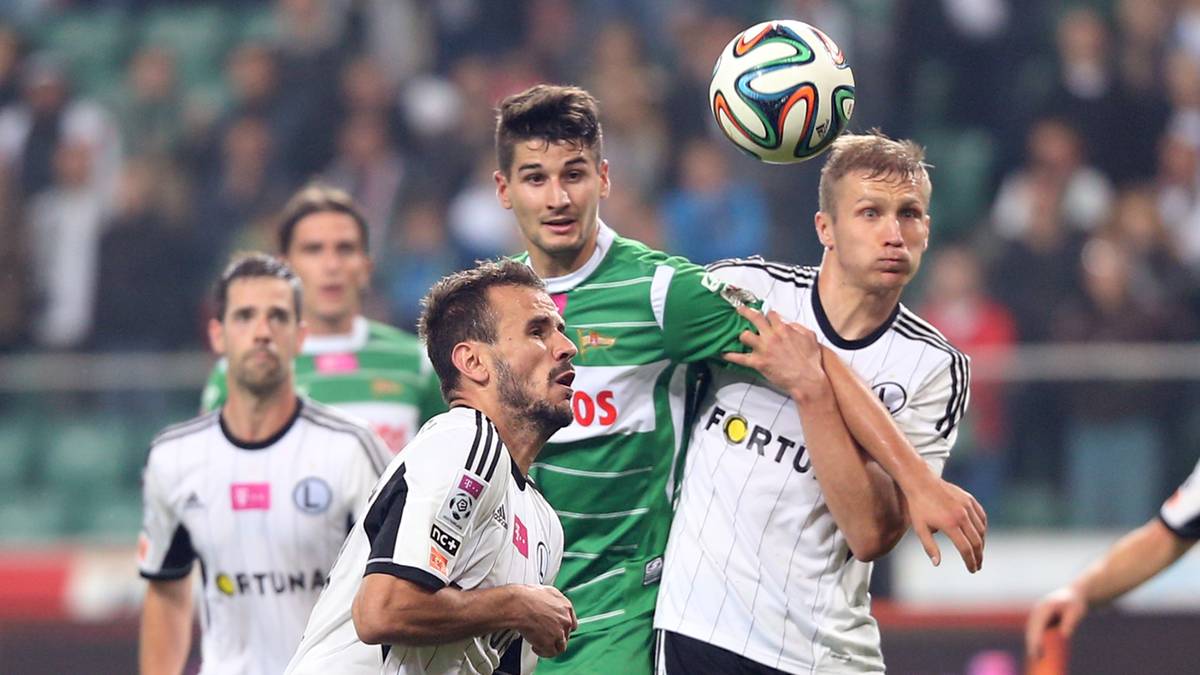 Piłkarze w białych strojach Legii Warszawa i zielonych strojach rywali walczą o piłkę.