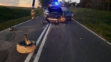 Miejsce wypadku drogowego z uszkodzonymi samochodami na drodze asfaltowej, z widocznym kołem od pojazdu i służbami ratunkowymi.