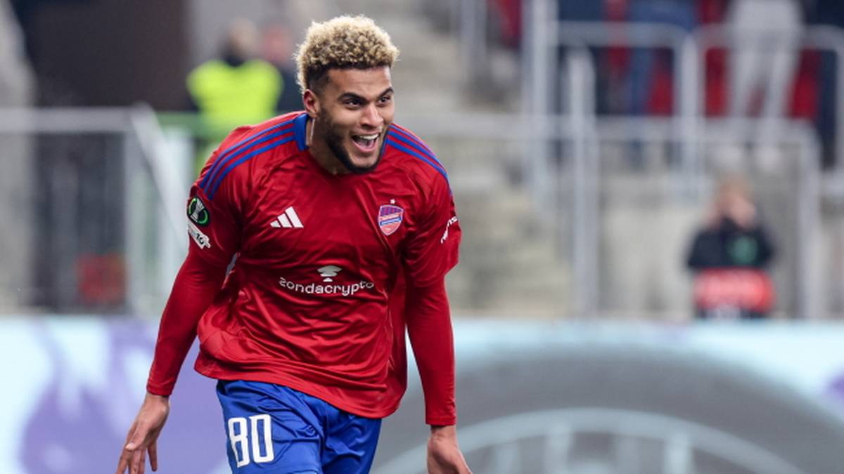 Piłkarz w czerwonej koszulce i niebieskich spodenkach z blond włosami i zarostem, z szeroko otwartymi ustami i uniesionymi ramionami, na tle stadionu.