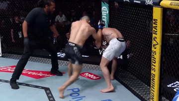 Dw&oacute;ch zawodnik&oacute;w MMA walczy w klatce, jeden z nich klinczuje przeciwnika pod siatką, obok stoi sędzia.